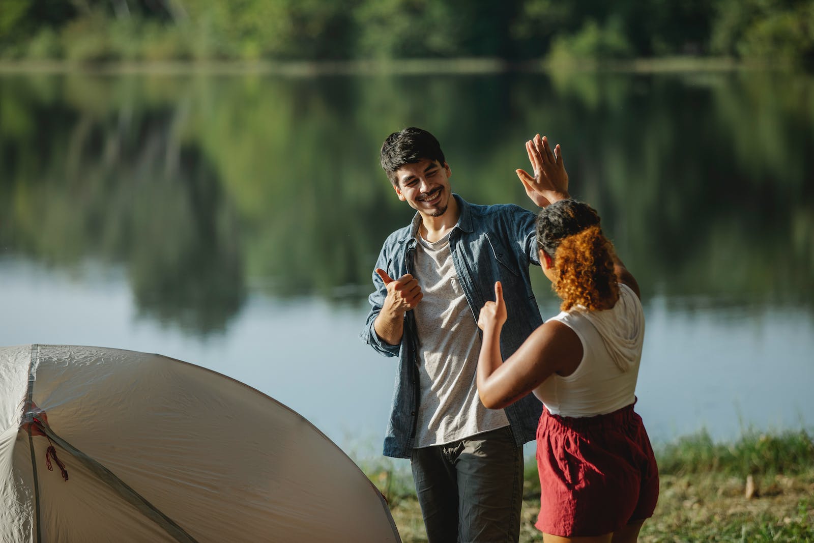 Glad male hiker with unrecognizable ethnic female beloved showing thumbs up and giving high five while looking at each other in campsite