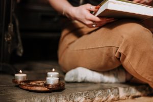 Unrecognizable mysterious female with book sitting in aged house near small burning candles placed on metal surface with spiritual symbol